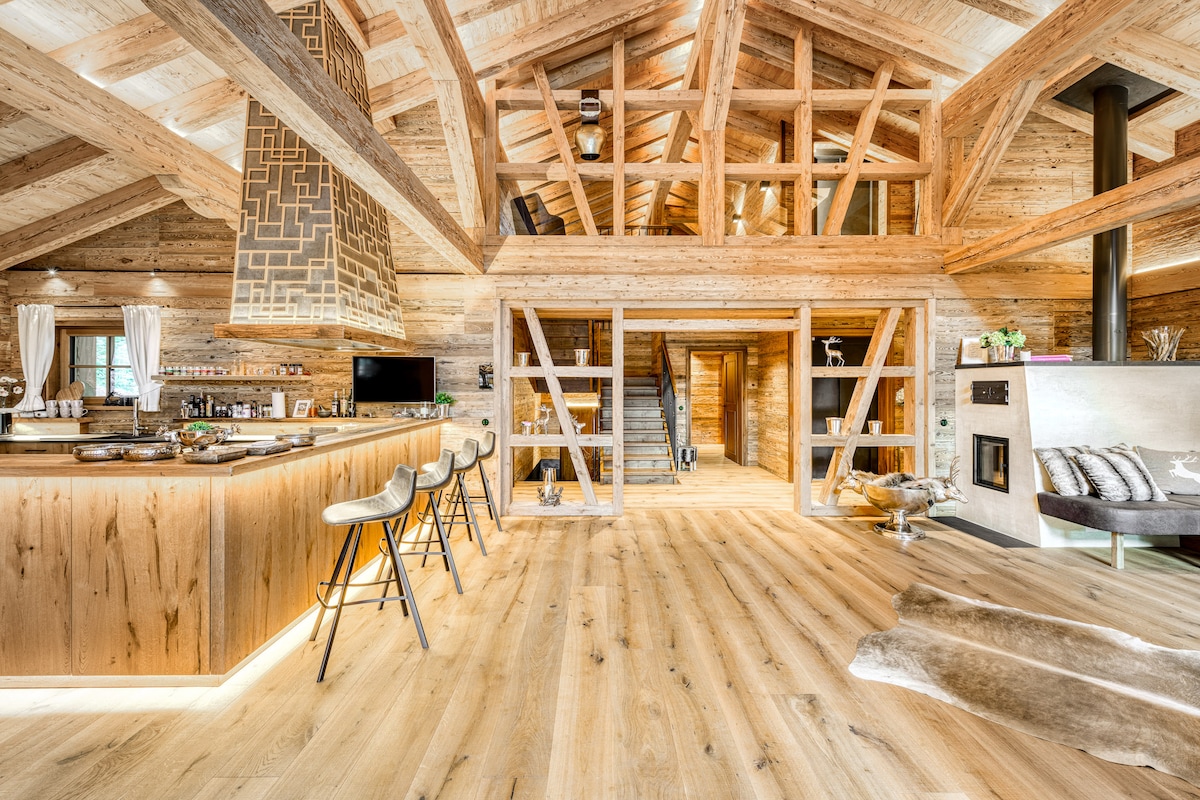 The interior of the chalet features a warm, wood-paneled design with an open and airy layout. A spacious kitchen island with bar stools provides a gathering spot, while shining lights highlight the grand timber rafters above. A staircase leads to additional living spaces, enhancing the flow of the area.