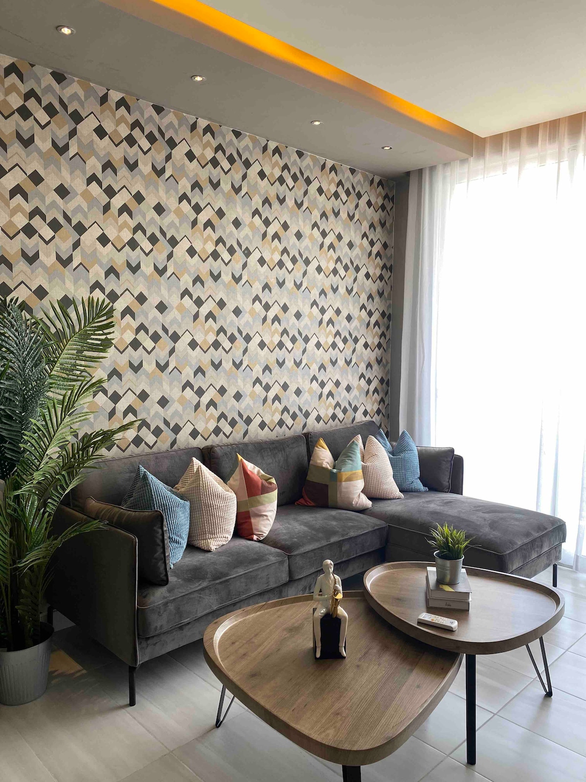 The living area features a modern gray sofa adorned with a variety of decorative pillows. A two-tier coffee table occupies the space in front of the sofa, complemented by a small potted plant. Natural light enters through sheer curtains, illuminating a textured geometric wall.