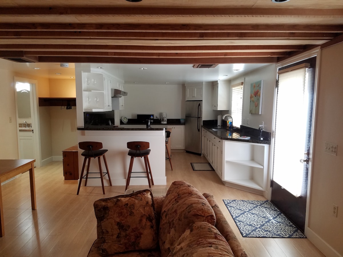 An open floor plan features a spacious living area with a comfortable couch and a dining table. The kitchen highlights granite countertops and is equipped with modern appliances. Two bar stools are placed at the kitchen counter, enhancing the communal space.