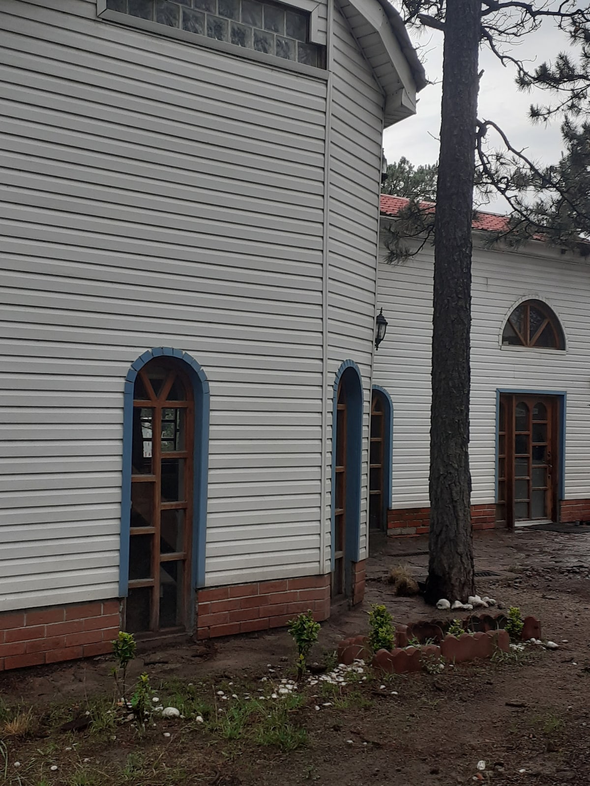 The exterior of a charming building features white wood paneling and arched windows outlined in blue. Two entrances, adorned with decorative details, are visible. Surrounding foliage includes small plants and mature pine trees, contributing to a natural, relaxing environment.