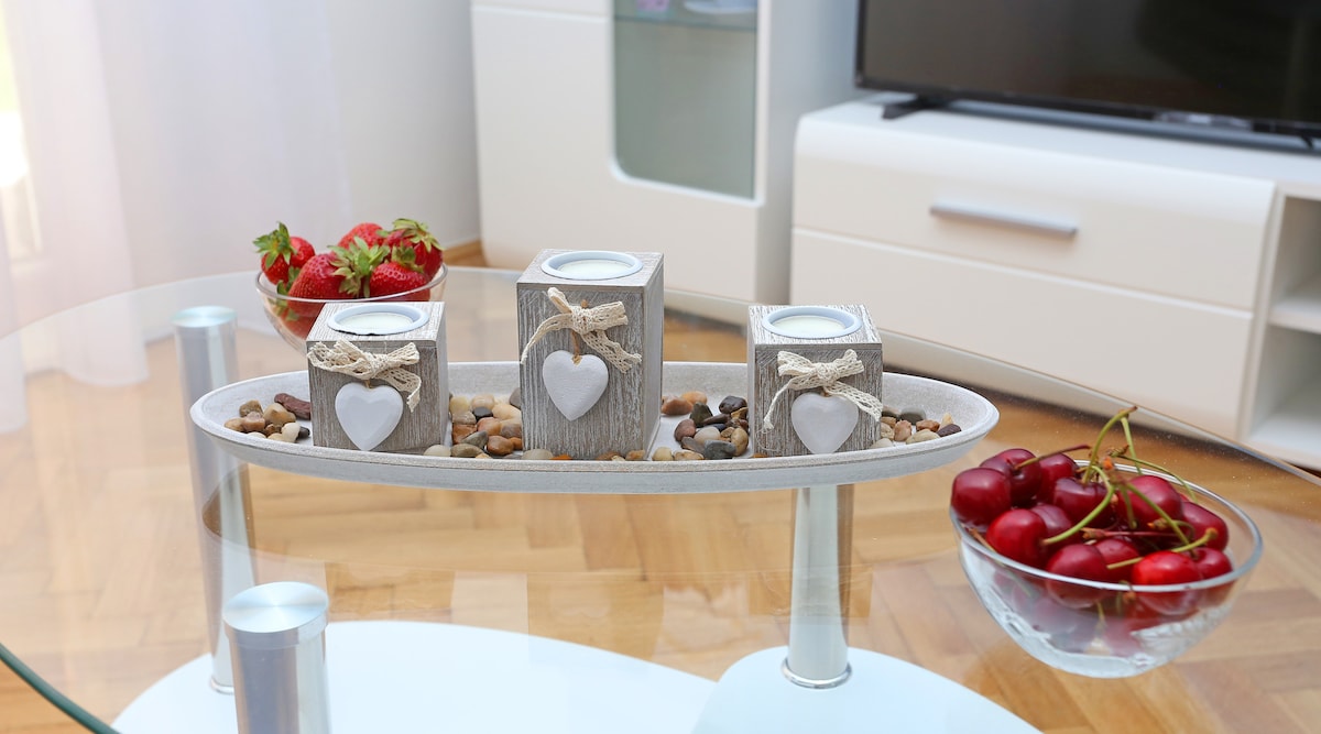 A modern coffee table is set with a round glass surface, featuring a decorative tray holding three wooden candle holders adorned with hearts. A bowl of fresh cherries and a small dish of strawberries are also displayed, adding a touch of colour and freshness.