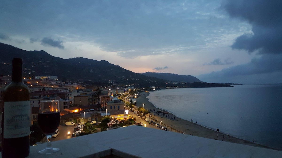 A view from a terrace captures the coastline at dusk, with soft lighting illuminating the beach. A wine bottle and glass are visible in the foreground, framing the expansive horizon where the sea meets the sky. The serene atmosphere invites relaxation.
