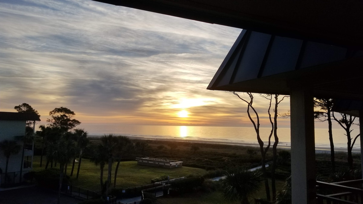 A sunset view is captured from the balcony, showcasing the golden sun descending over the ocean horizon. Silhouettes of palm trees and a grassy area are visible, creating a serene coastal atmosphere. The sky displays soft hues of orange, pink, and gray.
