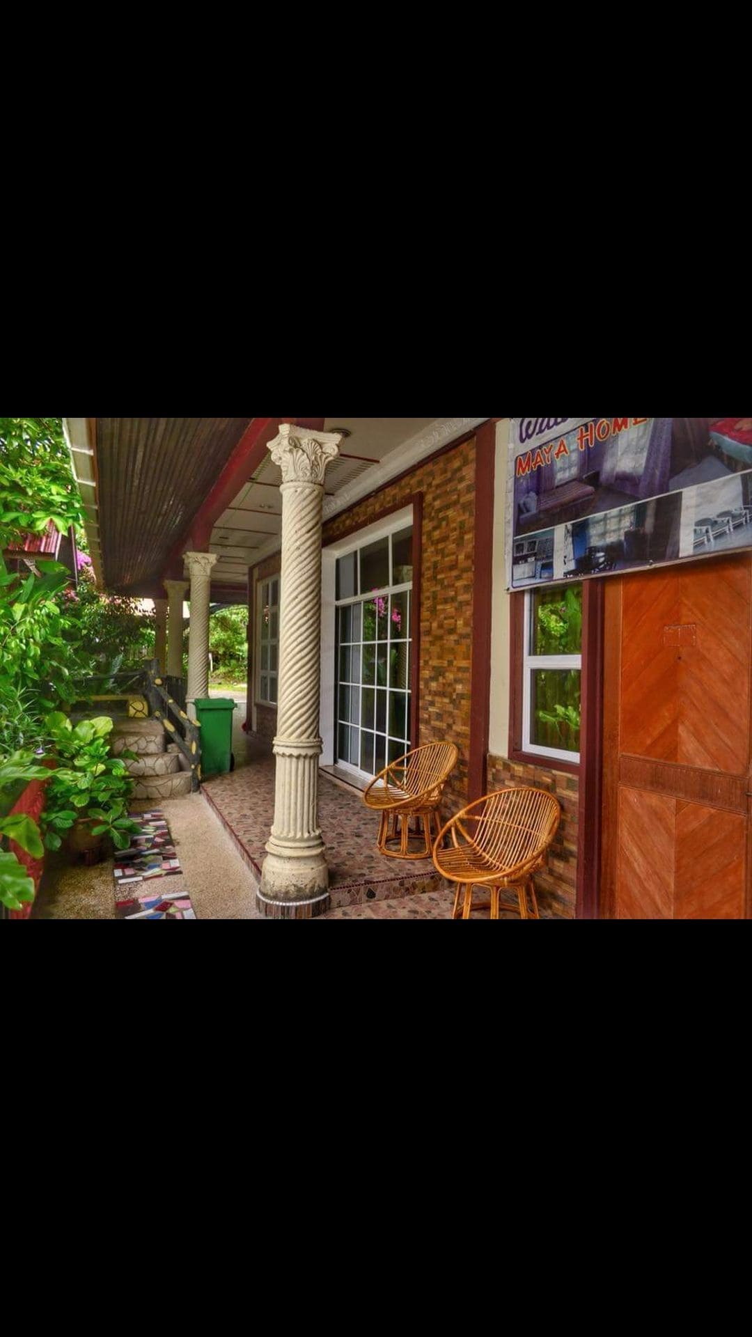 An inviting porch area features two woven chairs situated beside a decorative pillar. Lush greenery surrounds the space, and large windows provide a view into the interior. A well-maintained pathway leads to the entrance, enhancing the welcoming ambiance.