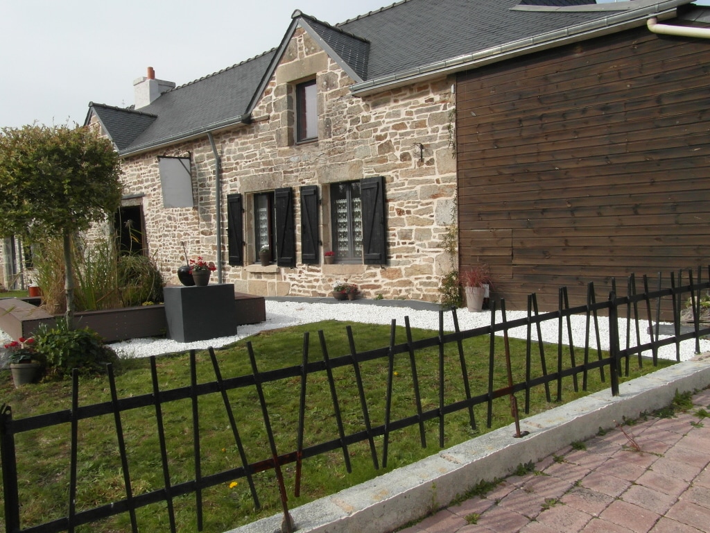 The exterior of the longère showcases a combination of traditional stone and modern wooden siding. A neatly maintained garden features a gravelled area with potted plants and flowers. A low fence complements the landscape, providing a boundary along the pathway leading to the entrance.