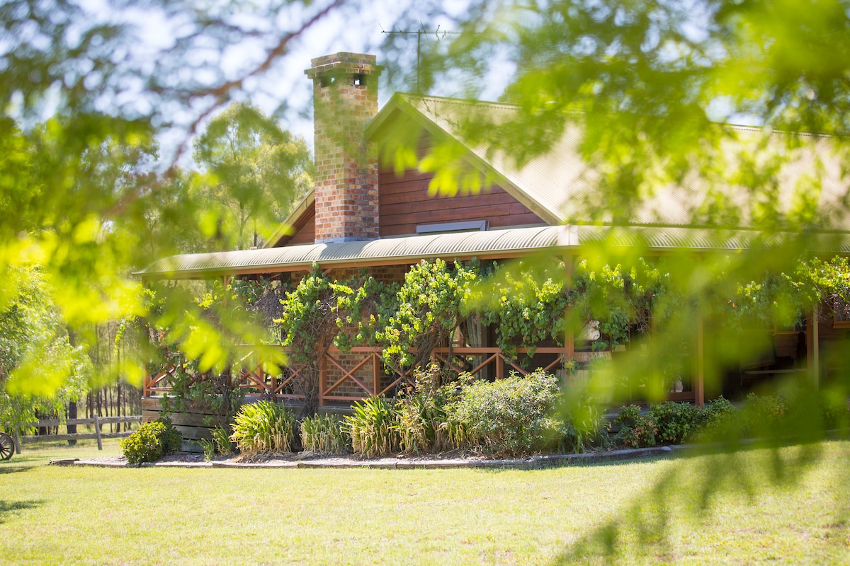 A timber cottage is nestled among lush greenery, showcasing a wraparound porch and a brick chimney. Sunlight filters through the leaves, highlighting the charming exterior and well-maintained garden that surrounds the property.