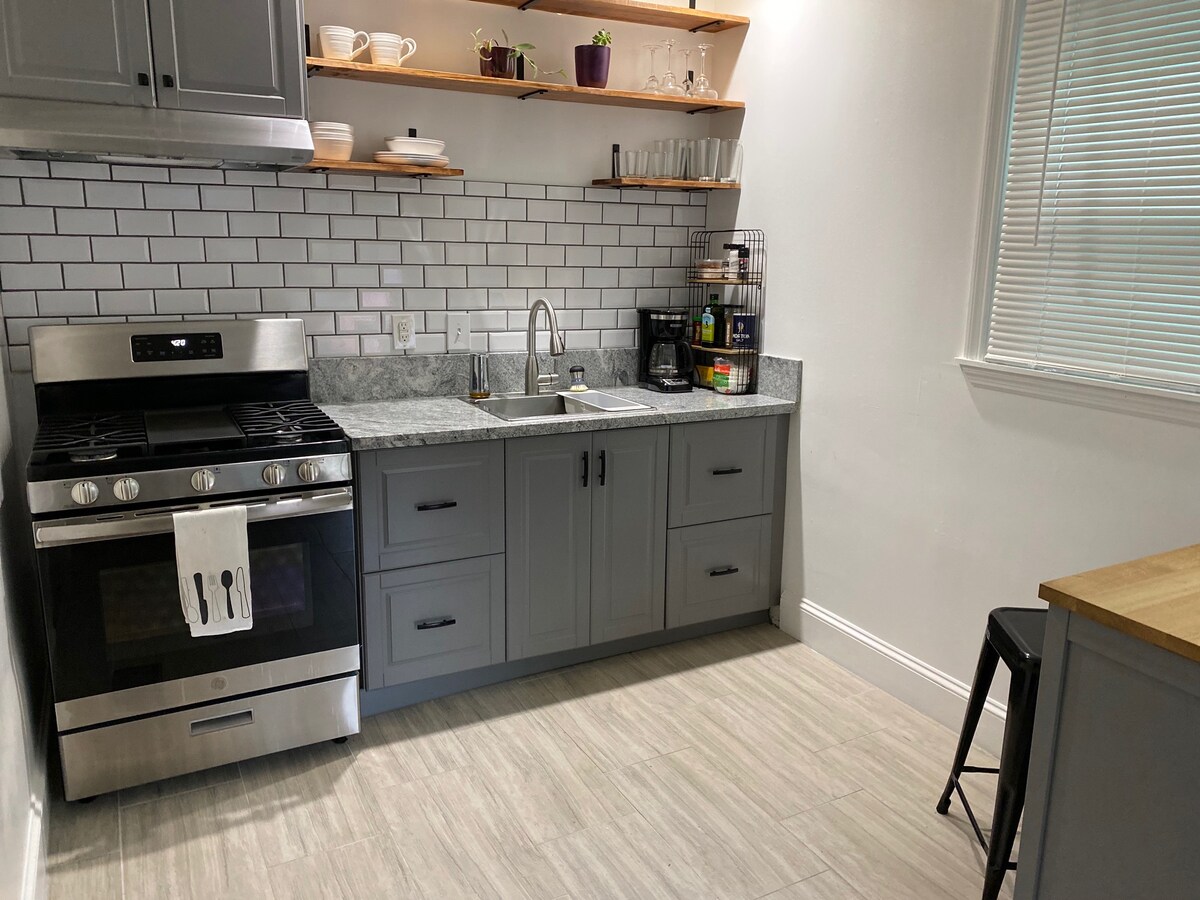 A modern kitchen is equipped with stainless steel appliances including a gas stove and refrigerator. Grey cabinetry complements a marble countertop. Open shelves display dishware and potted plants. A small wooden table with a black chair is positioned against the wall, providing a cozy dining spot.