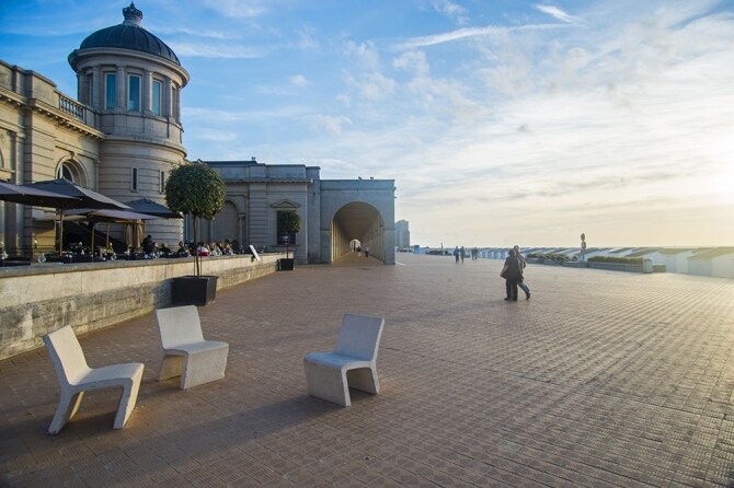 An open promenade is depicted, featuring modern white chairs arranged in a circle on warm-toned tiles. Sunlight casts a soft glow, illuminating the pathway and nearby café. In the background, people can be seen strolling leisurely, while elegant architecture creates a scenic backdrop.