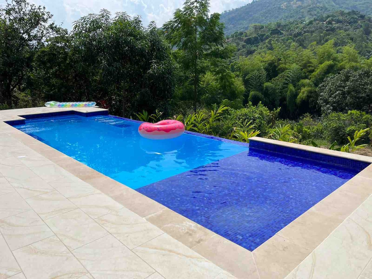 A serene swimming pool features clear blue water surrounded by lush greenery. A floating lounge in a bright color adds a touch of vibrancy. Stone tiles border the pool, leading to a gentle slope toward the natural landscape of mountains in the background.
