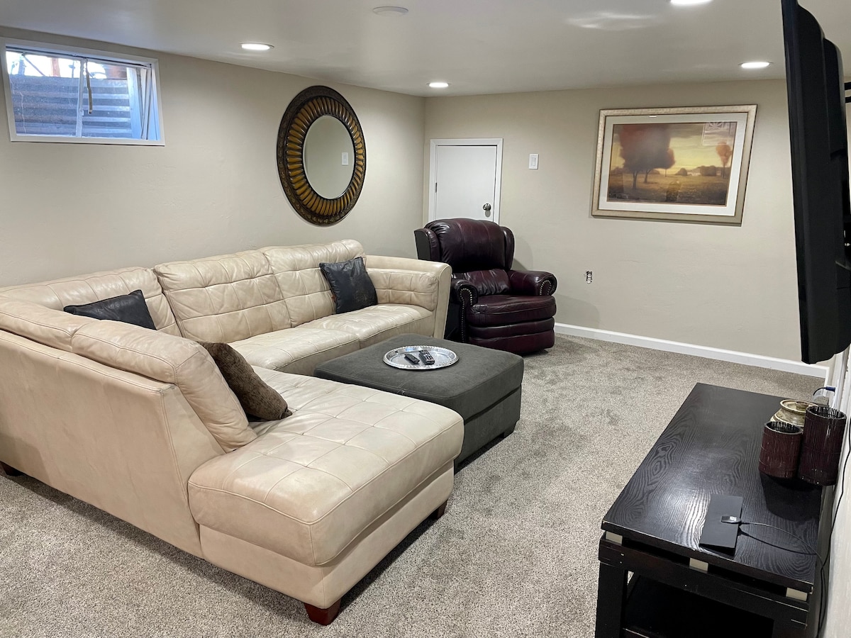 A spacious living area features a light-colored sectional sofa arranged around a square coffee table. A comfortable armchair is positioned nearby. The room is softly illuminated, with a large wall-mounted television and a decorative framed print on the wall.