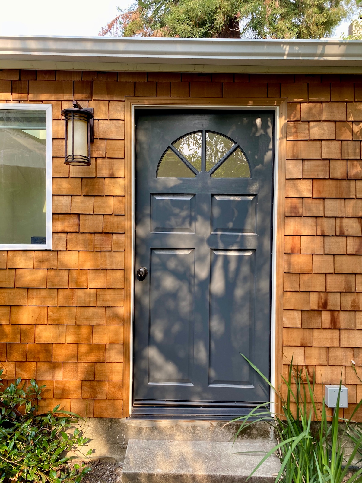 The entrance features a dark blue door with an arched window design, framed by a light wooden exterior with cedar shingles. Soft light filters through the nearby greenery, casting gentle shadows on the stone steps leading up to the door.