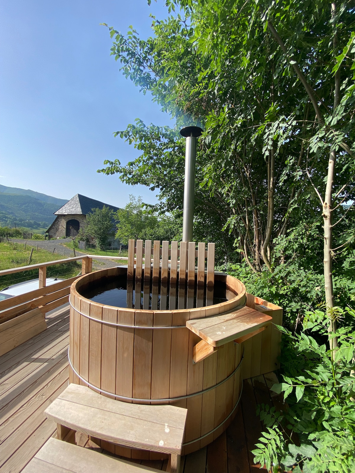 A wooden nordic bath is positioned on a spacious deck, surrounded by greenery. The bath features a wooden seating area and a chimney at the rear. Soft sunlight filters through the leaves, casting gentle shadows across the deck, while a scenic view of the countryside is visible in the background.