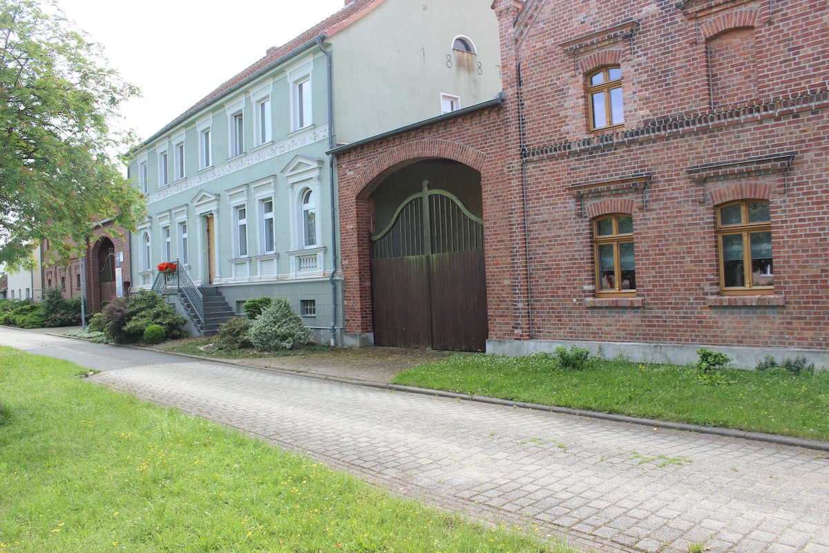 A historical farmhouse is visible, featuring a mix of green and red brick facades. The entrance includes a large wooden gate, surrounded by neatly maintained greenery and a stone pathway. Large windows provide ample natural light to the exterior spaces.