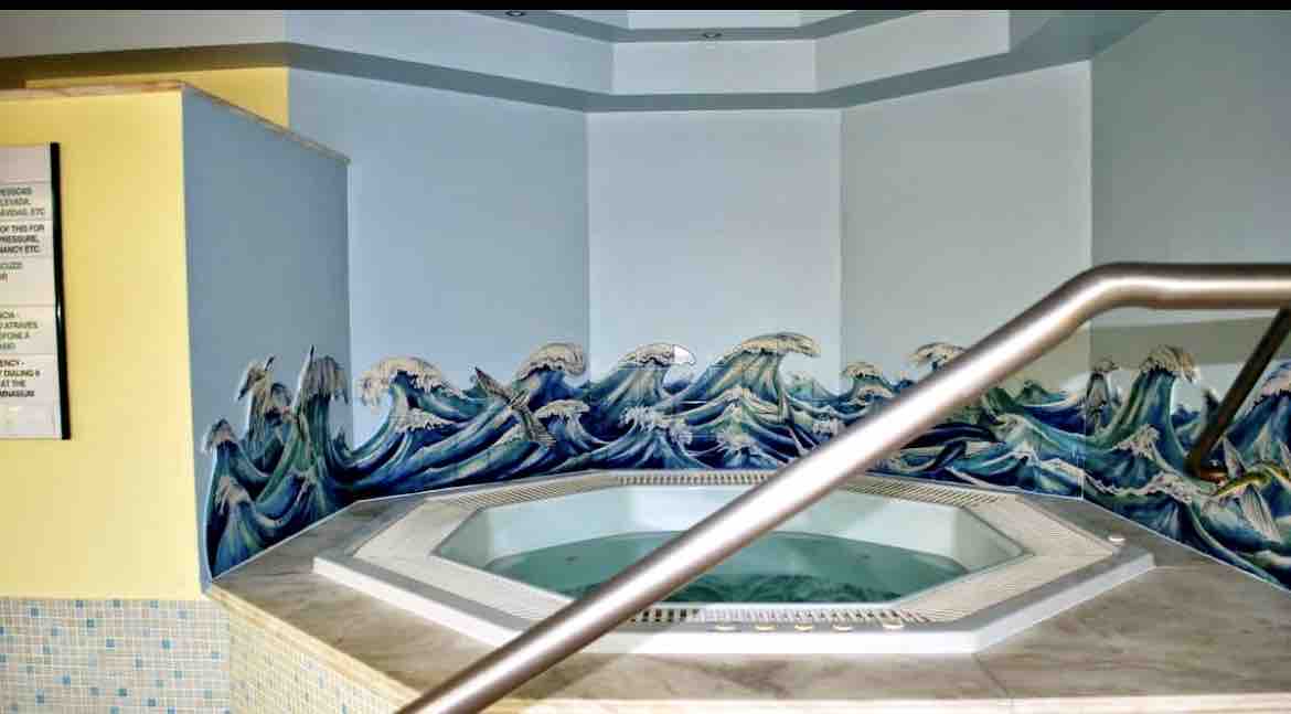 A modern jacuzzi is set in a bright room, featuring a colorful mural of waves along the walls. Natural light illuminates the space, highlighting the octagonal shape of the jacuzzi. A stainless steel railing provides support, enhancing the accessibility of the area.