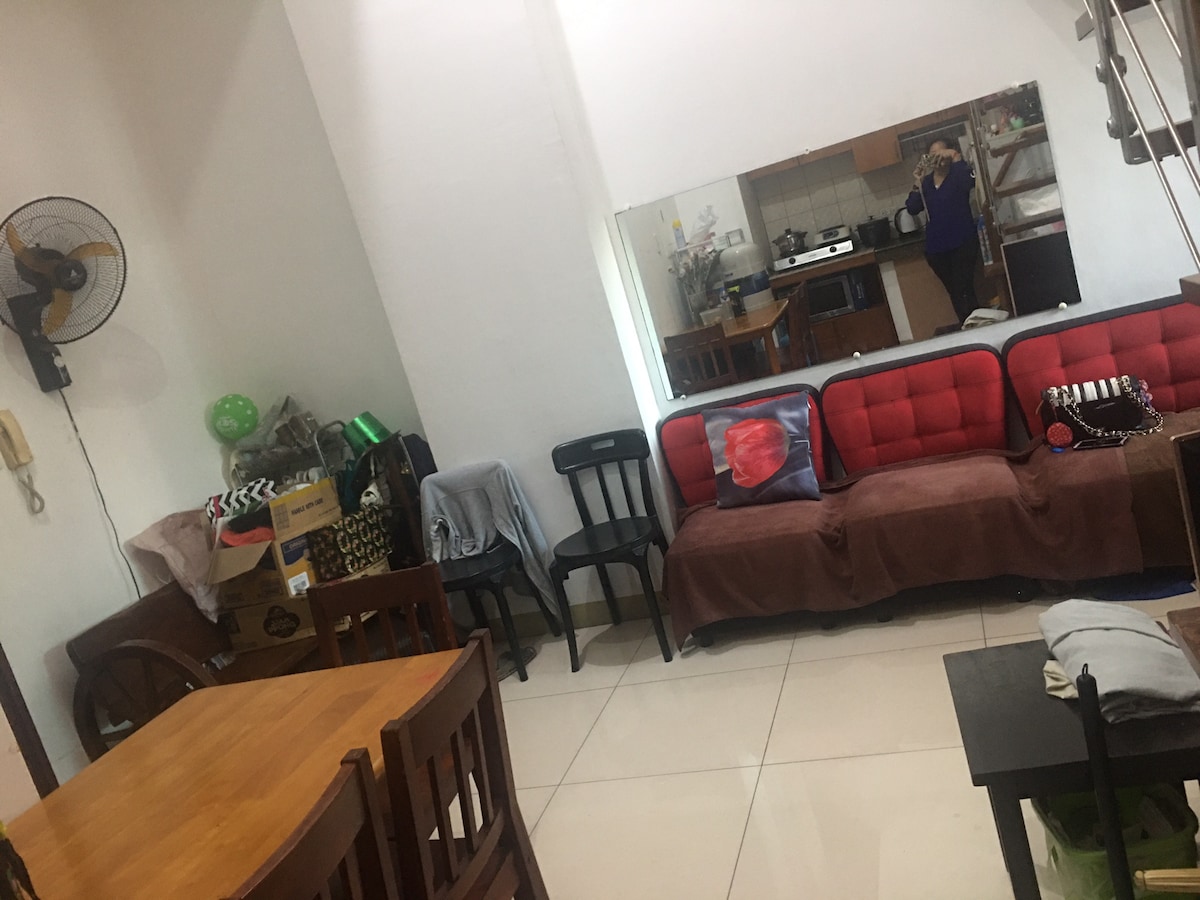 A living area features a mix of seating options, including a red couch and black chairs. A wooden dining table occupies one corner. Boxes and soft furnishings create a casual atmosphere. A large mirror reflects the space, while a fan is mounted on the wall.