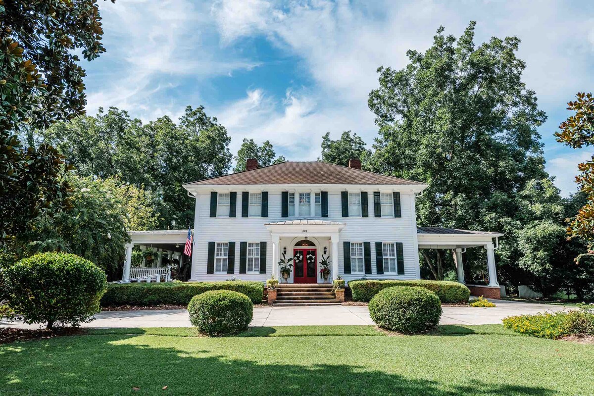 The historic home features a classic southern facade with white siding and dark green shutters. A welcoming red front door is framed by potted plants, while manicured shrubs line the pathway leading to the entrance. Tall trees provide shade and enhance the outdoor ambiance.