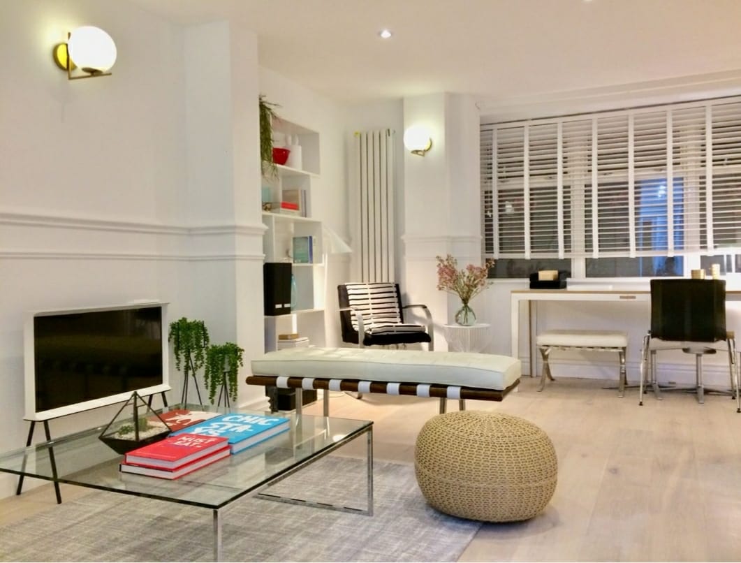 The living area showcases a minimalist design with a glass coffee table and a pouf. A sleek television sits against the wall, complemented by a striped chair. Natural light filters through the wooden blinds, highlighting the contemporary decor and greenery displayed on shelves.