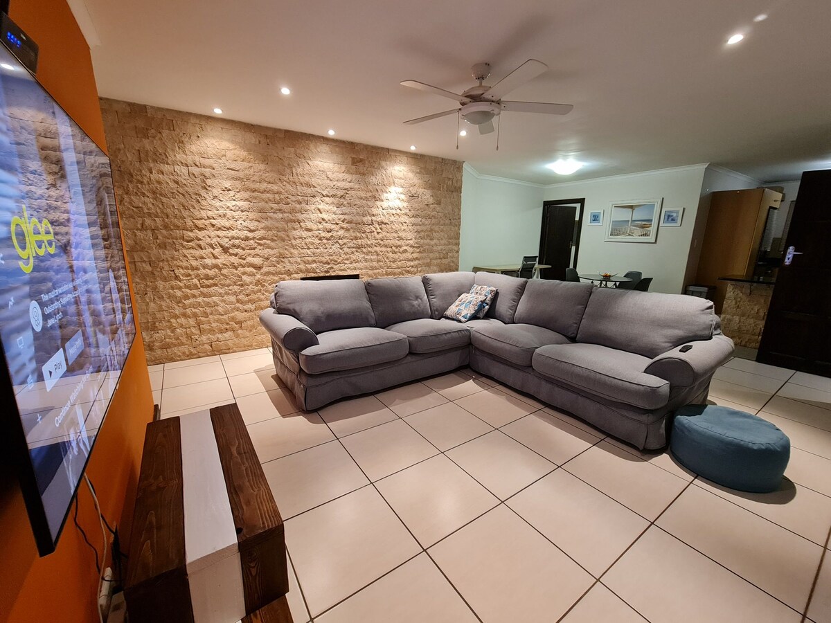A spacious living area is presented with a large sectional sofa positioned in front of a wall-mounted television. Soft lighting accentuates the textured feature wall. A circular pouf is placed nearby, and a dining area is subtly visible in the background.