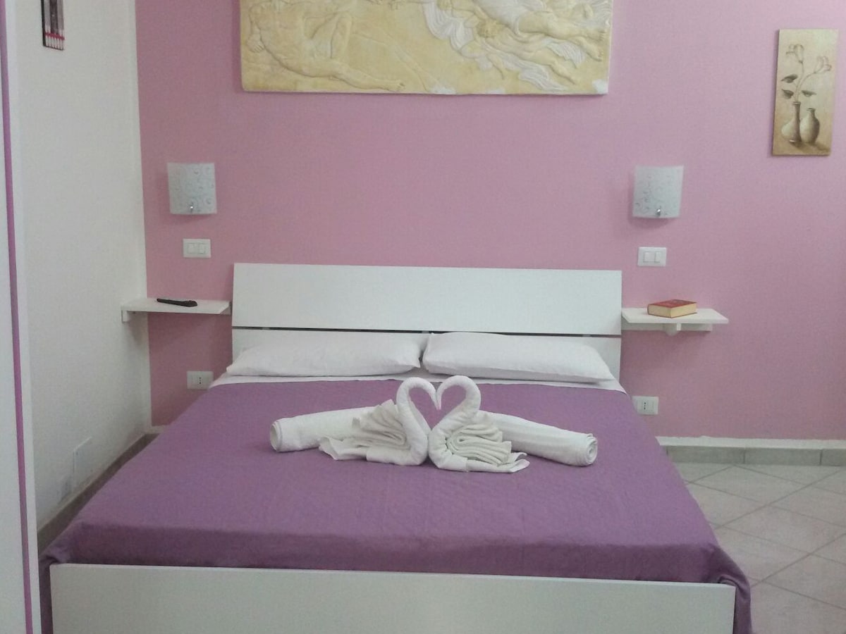 A sleeping area is furnished with a spacious bed, draped in purple bedding. Decorative towels are arranged in a heart shape on the covers. The walls are painted light pink, complemented by soft lighting from wall sconces. Two small shelves are situated beside the bed.