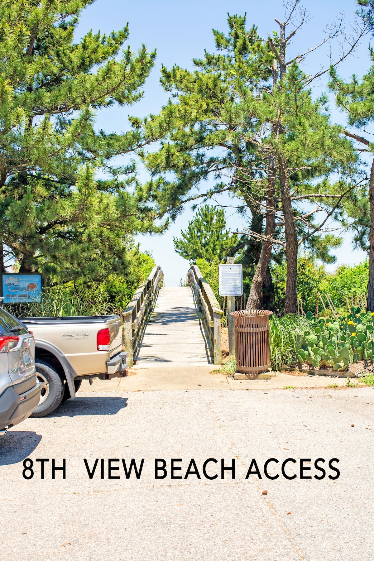 A wooden walkway leading to the beach is framed by tall pine trees. A sign indicating '8th View Beach Access' is visible near the end of the pathway. Nearby, a parking area with a parked vehicle can be seen, providing convenient access to the beach.