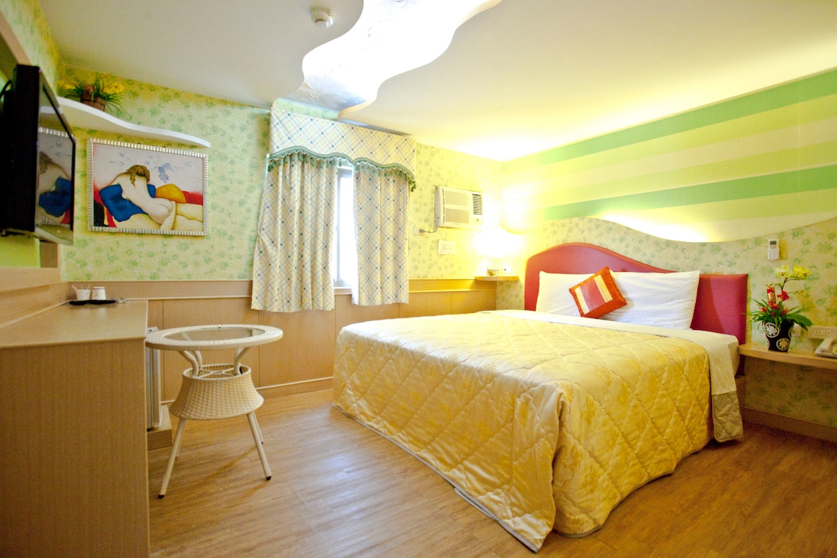 A bright room features a spacious bed dressed in a quilted gold comforter. Light green and floral wallpaper complements colorful artwork. A small round table and chairs are positioned nearby, with a window draped in patterned curtains offering natural light.
