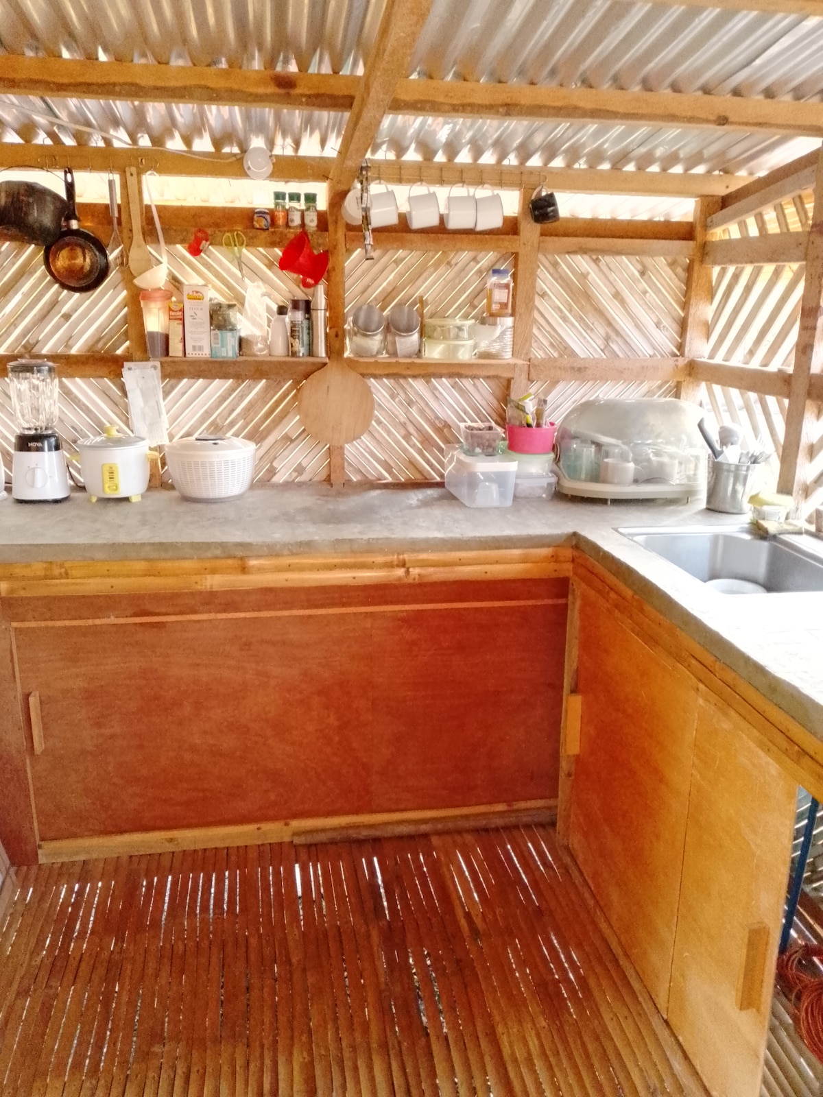 The kitchen area features wooden countertops and a double sink, with a variety of cooking utensils organized on hanging racks. Glass jars and containers are neatly arranged, while a blender and bowls are also visible. Natural light filters through the bamboo walls, creating a functional space.