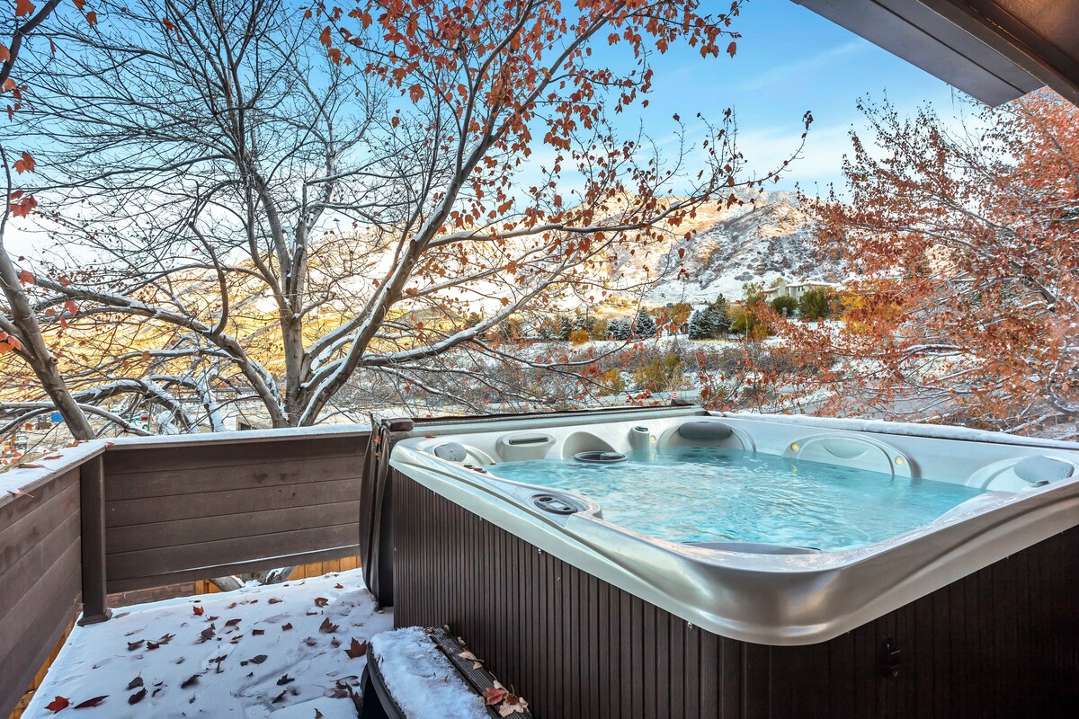 A hot tub is situated on a wooden balcony, surrounded by trees displaying autumn foliage. The serene water is framed by mountain views in the background, and a light dusting of snow is visible around the tub, enhancing the inviting outdoor space.