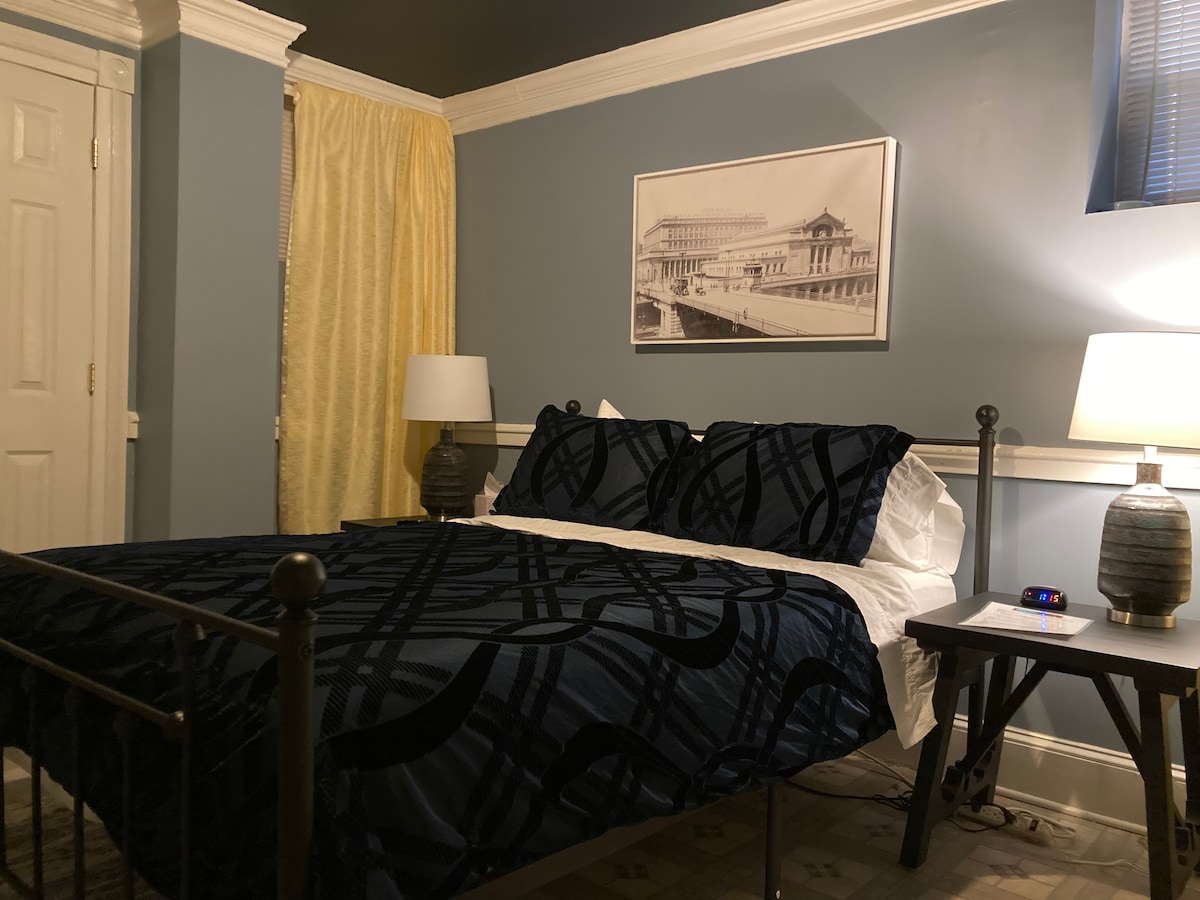 The room features a spacious bed dressed in a dark patterned blanket. A pair of bedside lamps illuminate the area, providing soft light. A framed black-and-white photograph adorns the wall above the bed, and yellow curtains add a touch of color to the room.
