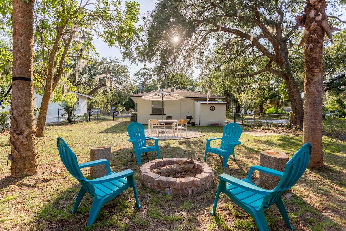 A charming backyard scene is captured, featuring a circular fire pit surrounded by four turquoise chairs. Sunlight filters through the trees, illuminating the grassy area. A table with chairs is positioned nearby, inviting outdoor dining and relaxation under the shade of the surrounding trees.