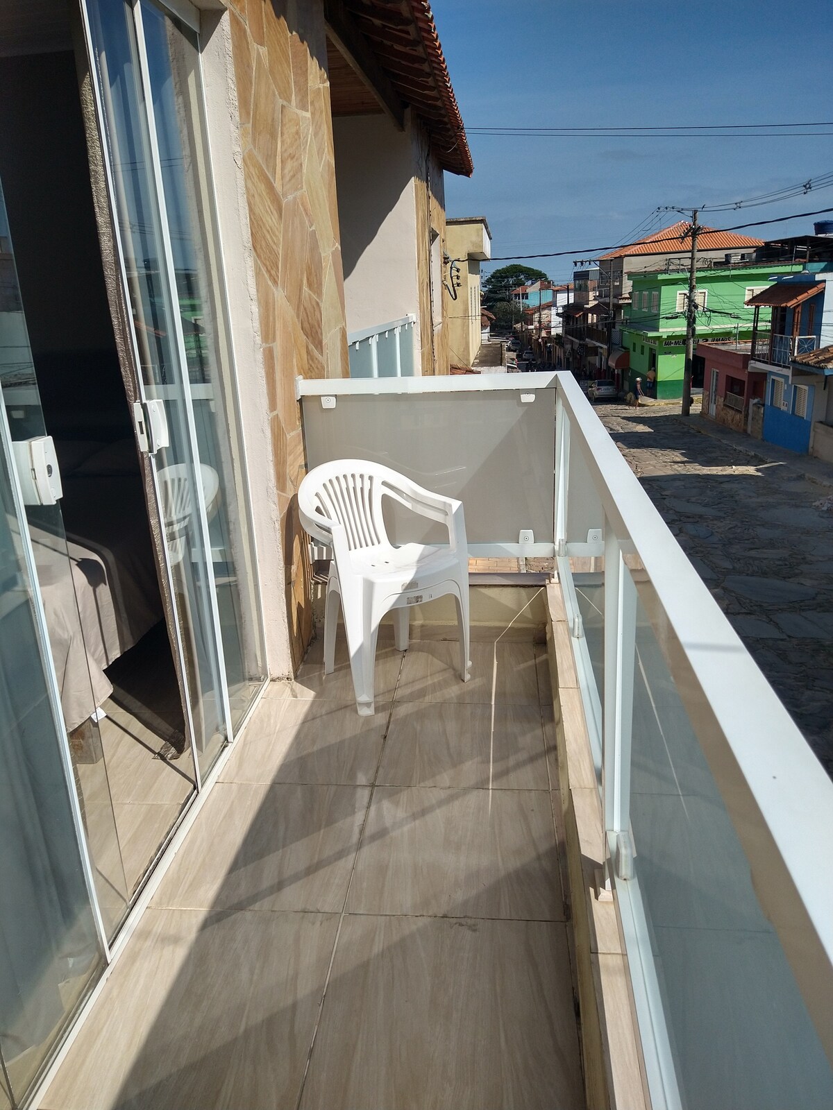 A private balcony is featured, with a lightweight plastic chair positioned beside glass railings. The floor is tiled and offers views of a street lined with colorful buildings. Natural light enhances the bright and airy feel of the space.