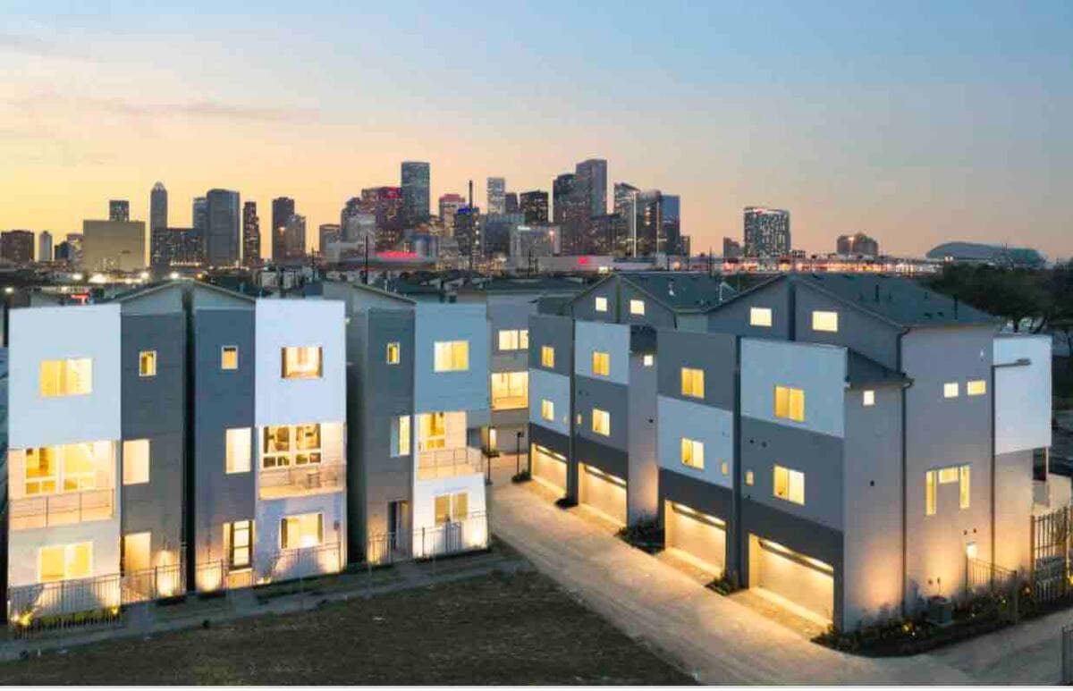 A modern multi-unit building is showcased at dusk, with soft outdoor lighting illuminating its facade. The skyline of a bustling city is visible in the background, featuring tall buildings against a pastel-colored sky.