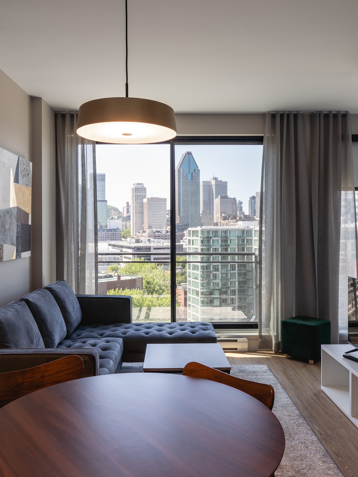A modern living space features a comfortable sectional sofa with a textured fabric. Large windows allow for natural light and provide a view of the downtown skyline. A wooden dining table and a green accent stool are visible, creating a functional and inviting area.