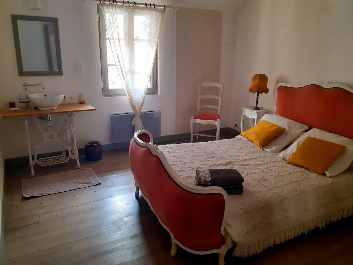 A charming bedroom features a spacious bed with soft bedding and decorative cushions. Natural light filters through sheer curtains over a window. A vintage-style chair, a wooden washstand, and a bedside lamp enhance the inviting space, contributing to a restful ambiance.