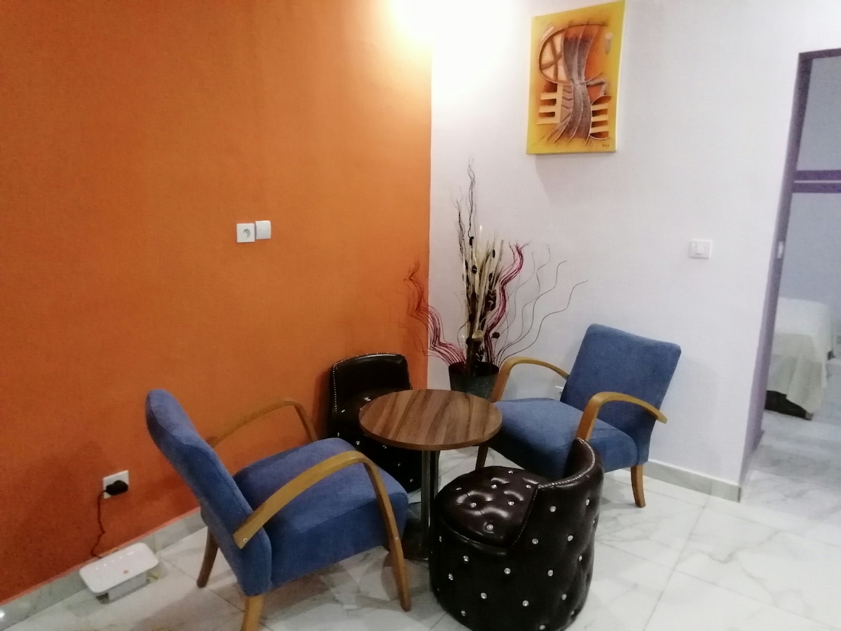 A cozy seating area features two blue armchairs and a round wooden table with a patterned design. A decorative plant in a vase adds a touch of elegance to the space. The walls are painted in orange and light purple, enhancing the atmosphere.