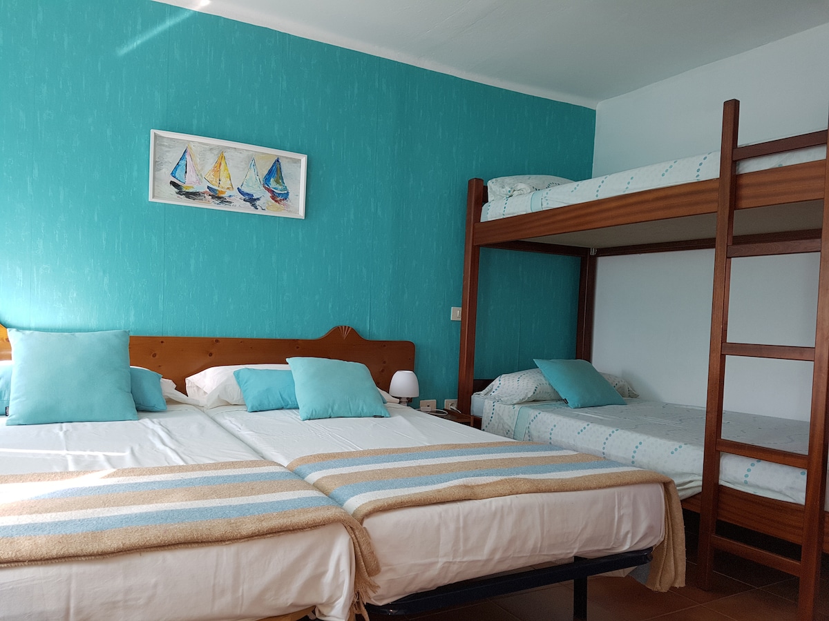 A comfortable private room is shown, featuring a spacious double bed alongside a bunk bed. Light blue accents are seen on the walls and bedding. Simple decor is complemented by a piece of nautical artwork above the beds.