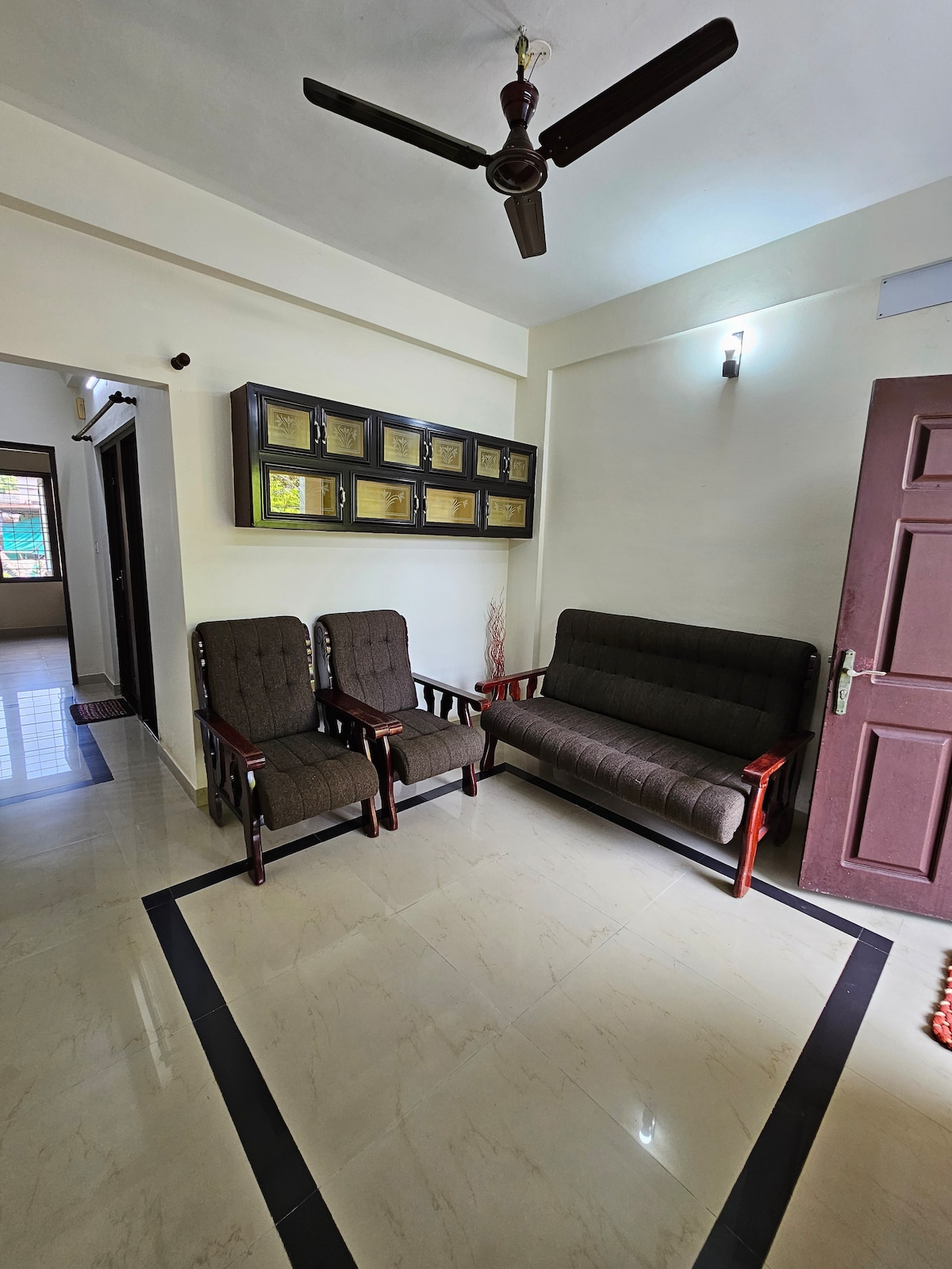 A well-lit sitting area features a comfortable brown sofa and two armchairs, arranged around a central space. A ceiling fan hovers above, and a decorative wall adorned with framed items adds interest to the neutral-toned room.