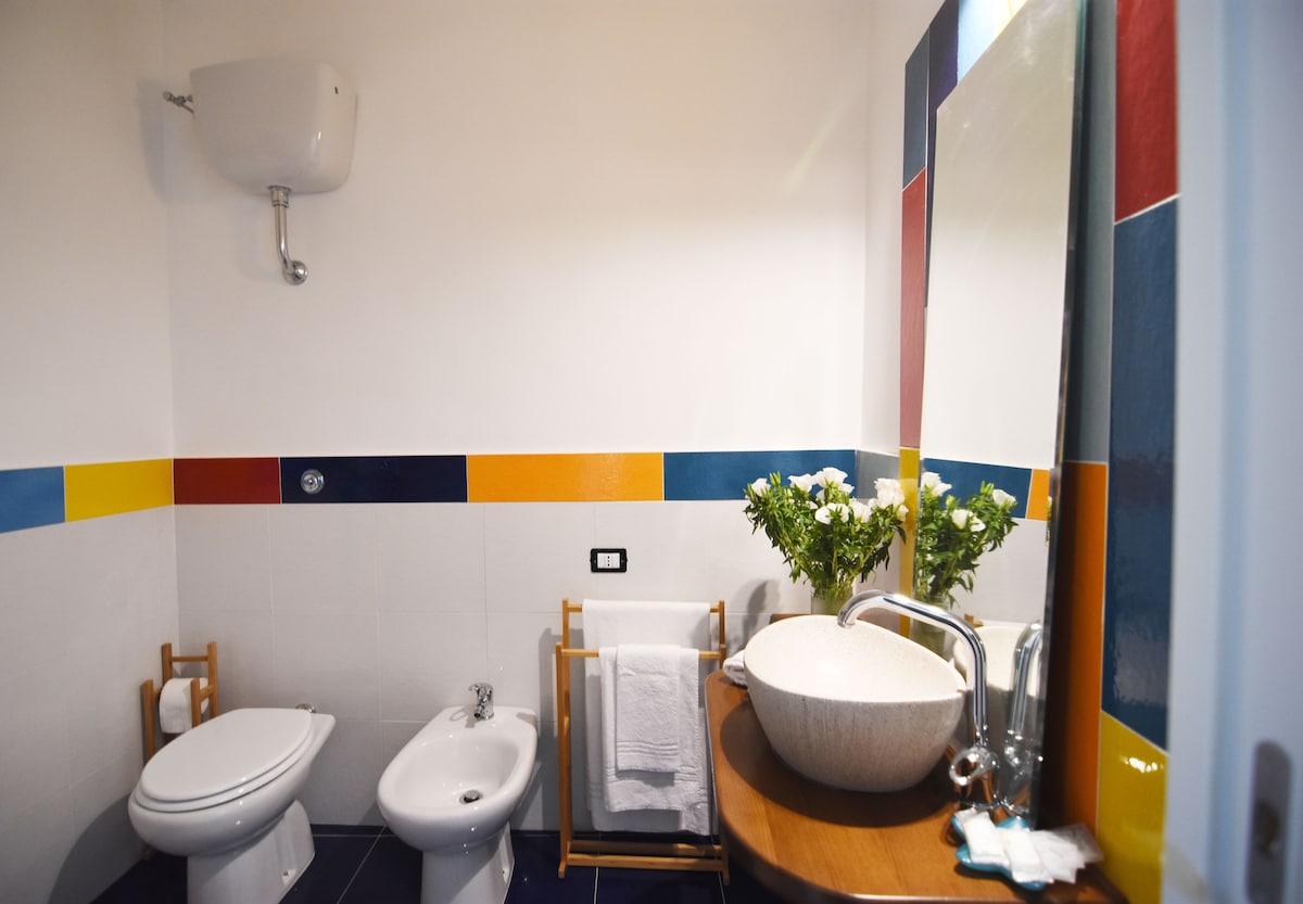 A modern bathroom is presented with colorful accents in the tiles. A round basin sits on a wooden countertop beside fresh flowers. A bidet and a toilet are positioned against the wall, and neatly hung towels are visible on a wooden rack.