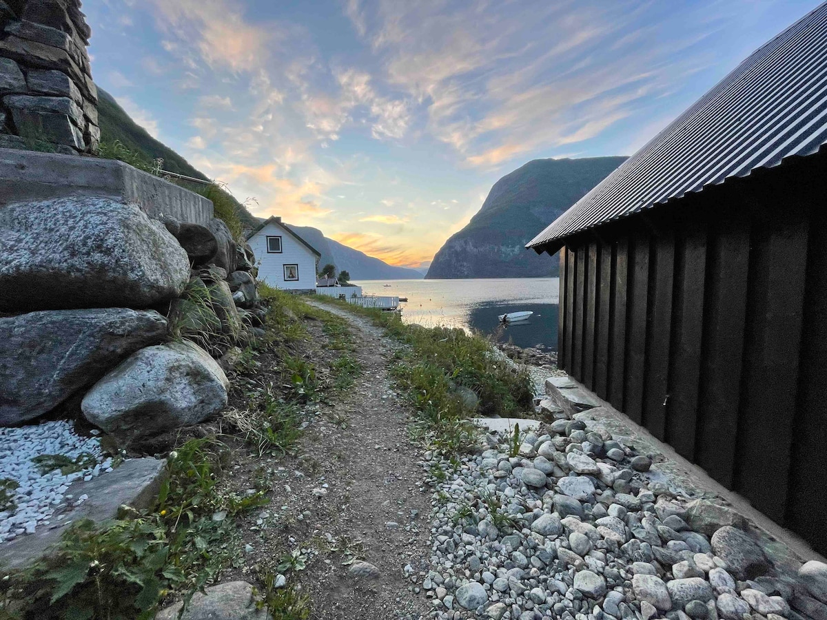 A winding gravel path leads toward the fjord, flanked by smooth stones and greenery. On one side, a white house is visible, while a rustic black boathouse is positioned nearby. The sky displays soft hues as the sun sets, reflecting tranquility over the water.