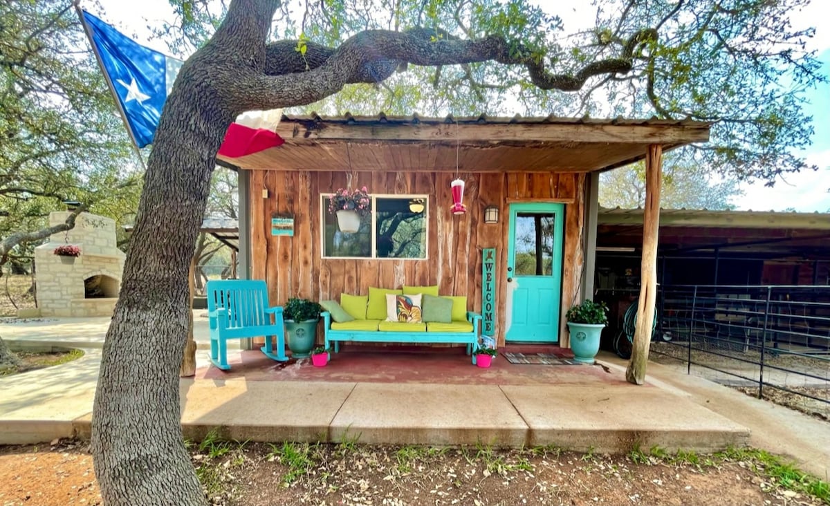 A charming cabin entrance is framed by a large oak tree, showcasing a bright blue door and vibrant seating. The welcoming porch features colorful cushions and potted plants, with a Texas flag gently fluttering nearby, creating an inviting outdoor space.