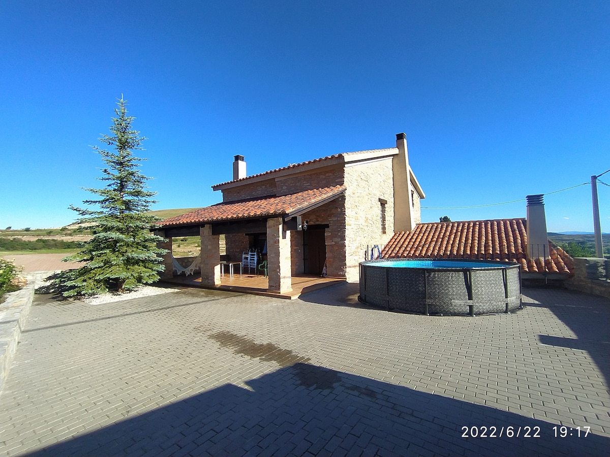 A rustic stone house is presented against a clear blue sky, featuring a terracotta-tiled roof and a spacious outdoor area. A circular hot tub is positioned next to the house, while a coniferous tree adds a natural touch to the landscape.