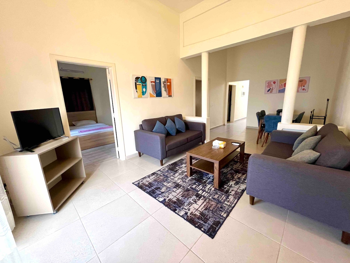 The living area is furnished with two gray sofas and a wooden coffee table, complemented by a decorative rug. A television is positioned on a cabinet against the wall. The space features tall ceilings and is illuminated by natural light, creating an open and airy atmosphere.