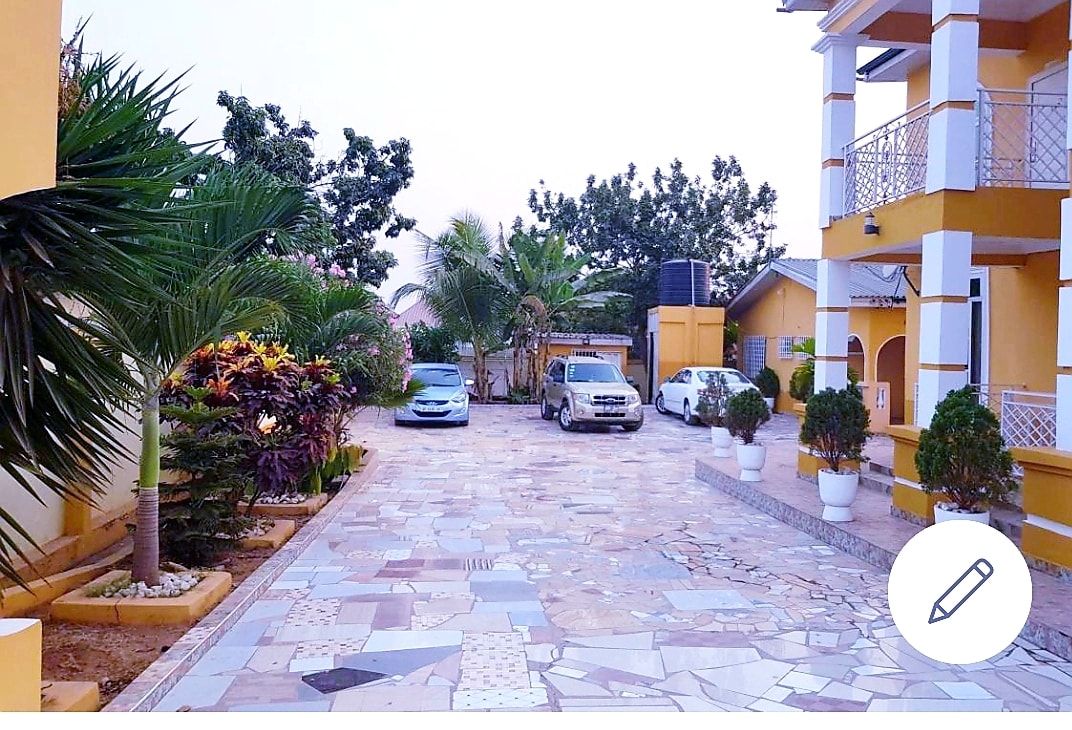The exterior courtyard showcases a well-maintained stone pathway surrounded by vibrant greenery and carefully placed decorative plants. Several parked cars are visible, and a warm yellow building stands in the background, providing a welcoming entrance to the compound.