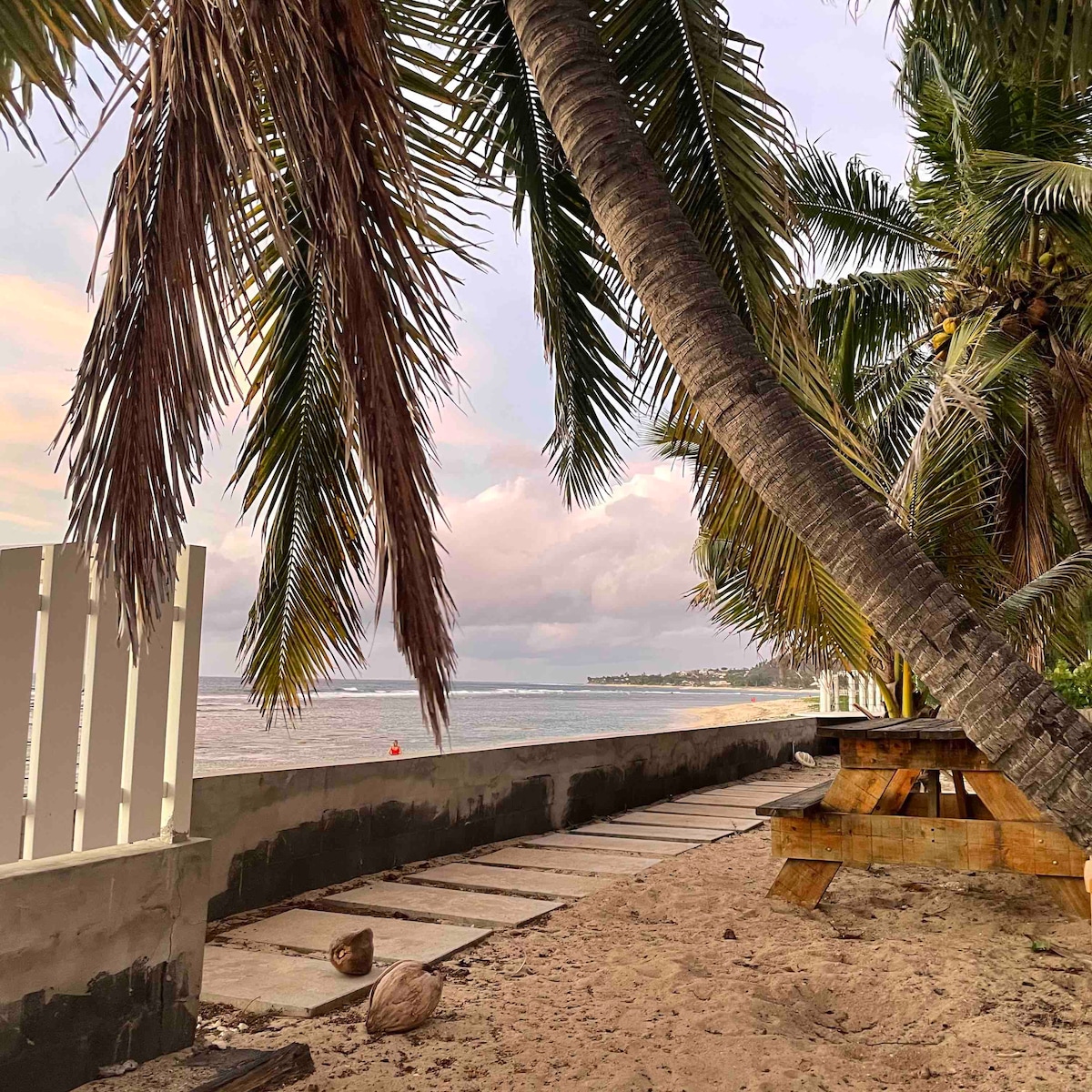 Palm trees frame a sandy area leading to the beach, where gentle waves can be seen in the distance. A wooden picnic table is positioned on the sand, providing a space for relaxation. The overall mood is tranquil, with soft colors reflecting off the water.