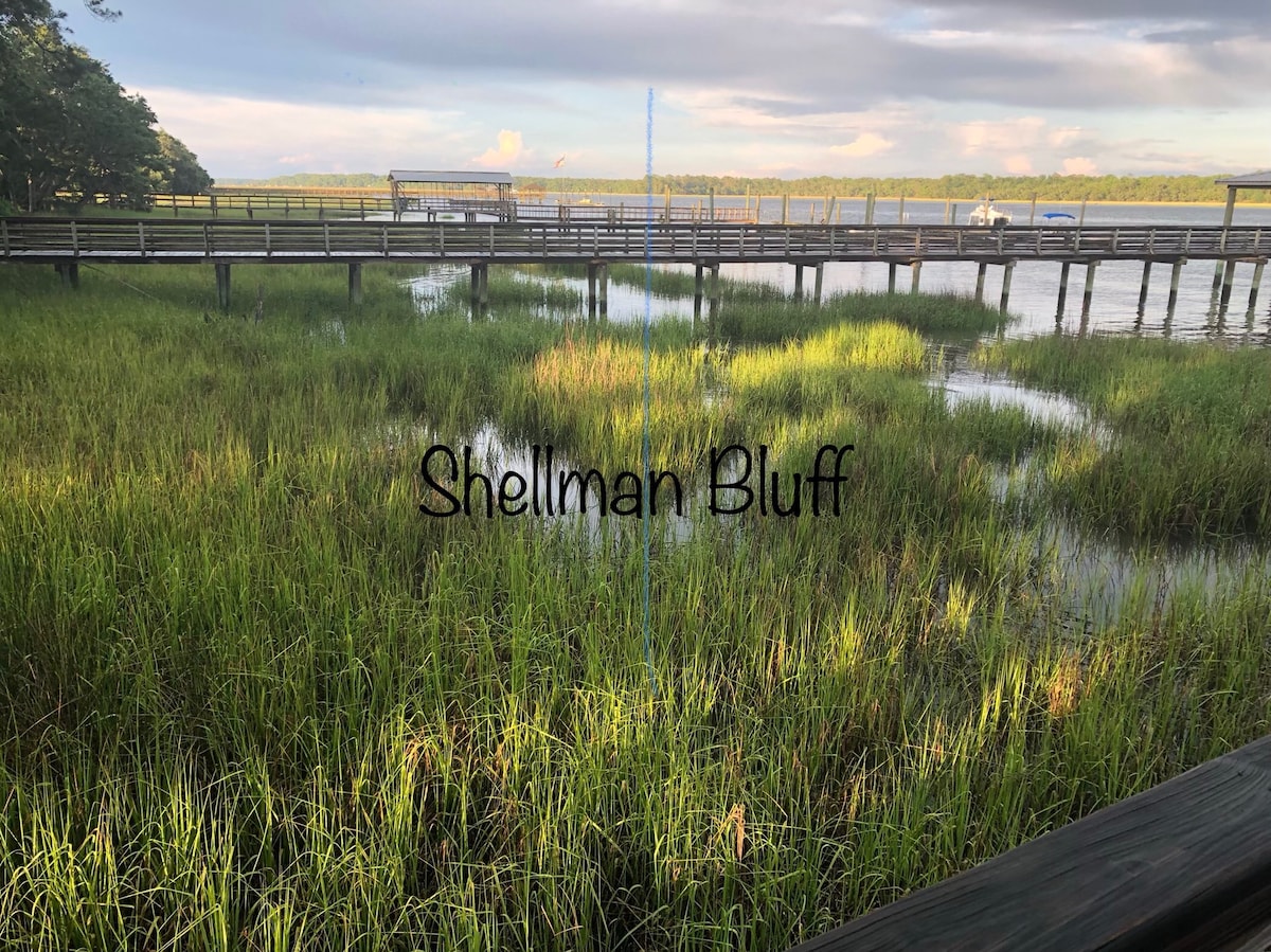 Lush green marsh grasses extend across the shallow water, creating a serene landscape. A wooden pier runs parallel to the water's edge, providing a view of the calm shoreline. The sky above showcases soft clouds against a fading light, enhancing the tranquil setting.