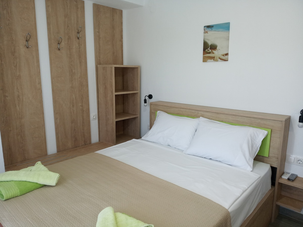 A spacious bedroom features a large bed with white linens and soft green accents. Simple wooden furniture includes a shelf and hooks for convenience. Natural light illuminates the room, enhancing its calming atmosphere.