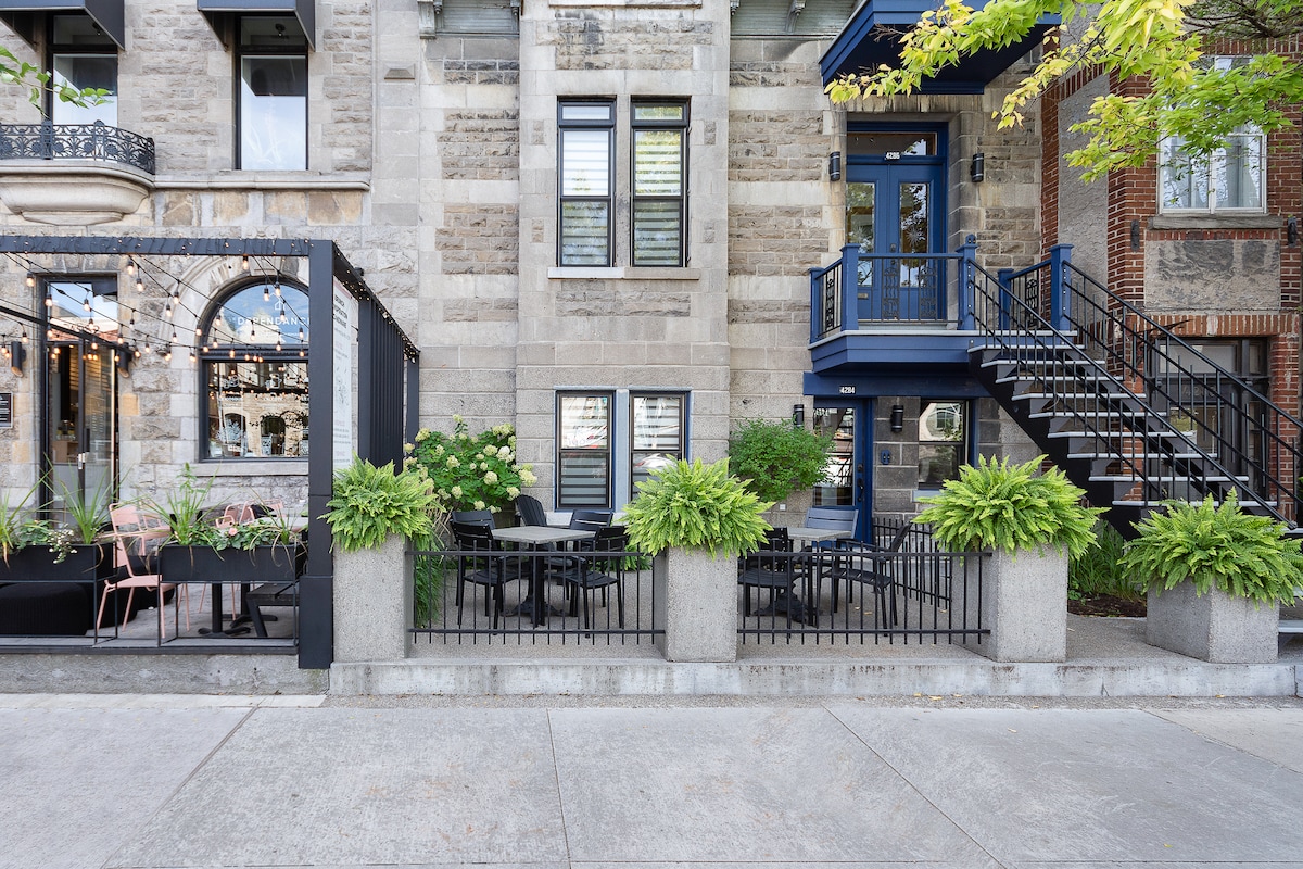 A charming outdoor terrace is visible, featuring elegant black furniture surrounded by lush greenery and decorative planters. The stone façade of the building provides a historical backdrop, complemented by blue accents on the entrance and stairs, enhancing the inviting atmosphere of the space.