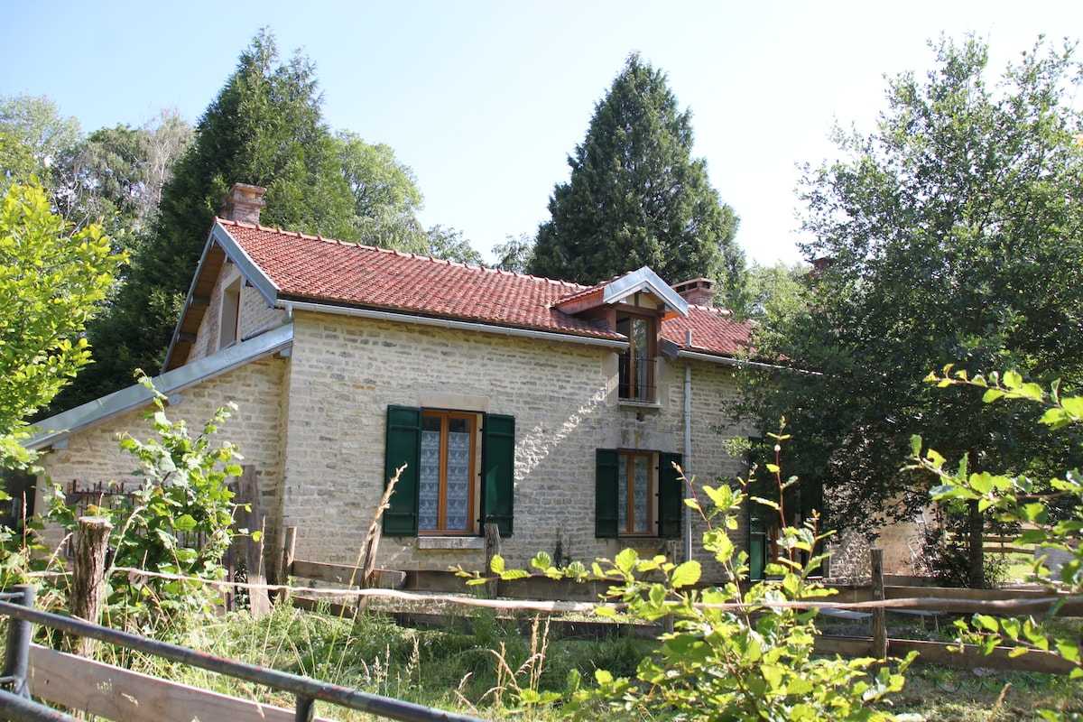 The exterior of a charming forest house is framed by lush greenery. The stone façade displays wooden shutters and a red-tiled roof, complemented by a small balcony. A fence surrounds the property, highlighting the serene natural setting.