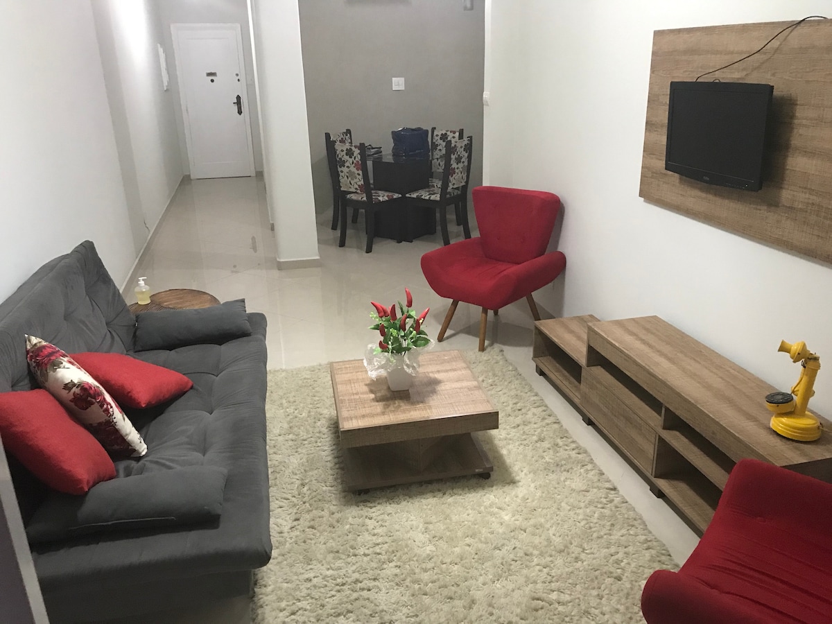 A modern living area features a soft, beige rug covering the polished floor. A dark gray sofa, adorned with red cushions, sits alongside a vibrant red armchair. A wooden coffee table holds a potted plant, while a compact entertainment unit with a television is mounted on the wall.