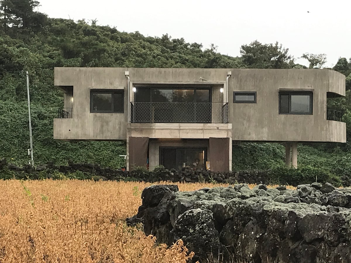 The modern structure is set against a backdrop of lush greenery and a rustic stone wall. Its unique design features large windows and a balcony, providing a view of the surrounding landscape. The concrete exterior integrates with the natural surroundings.