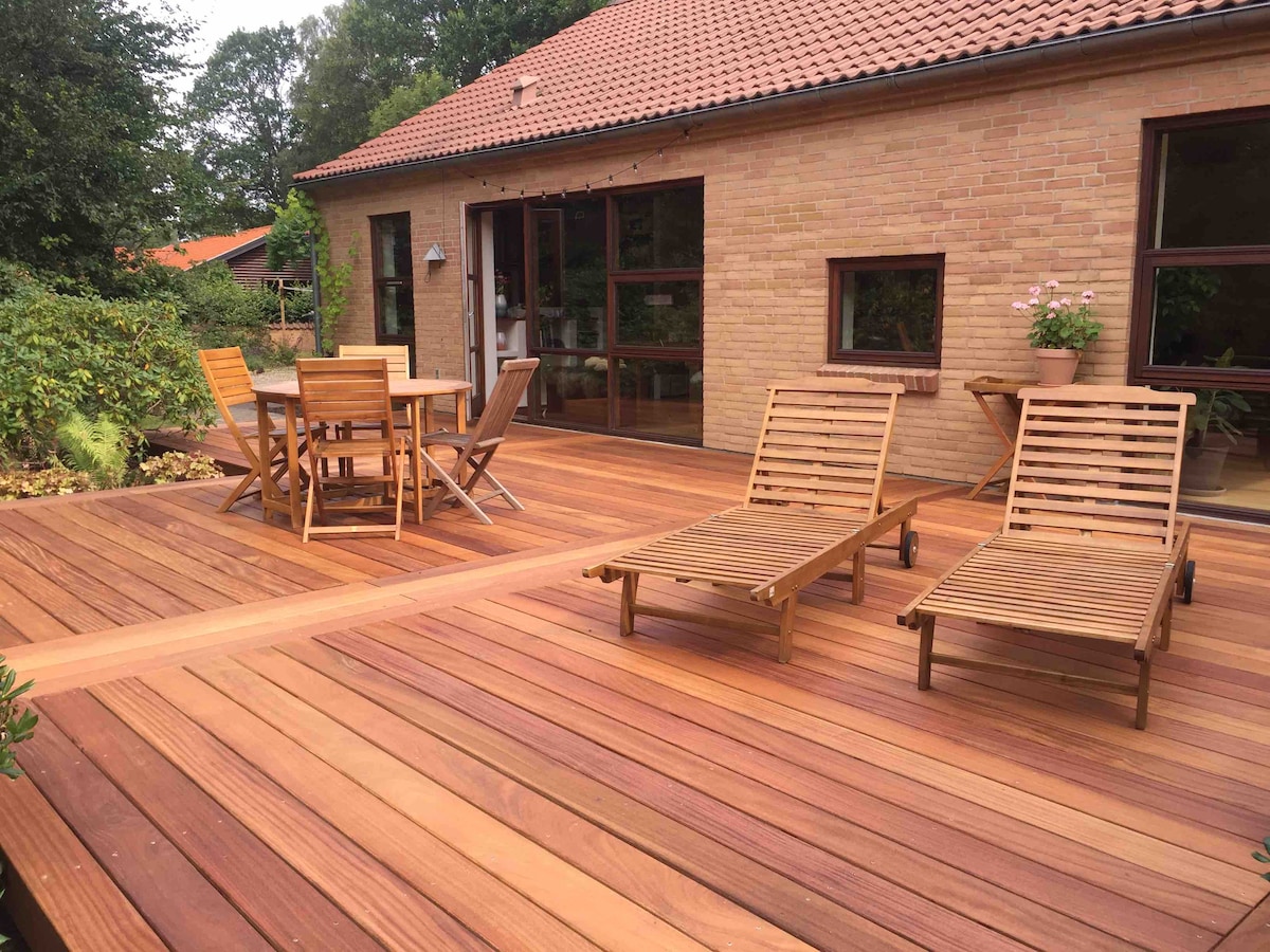 A wooden terrace features a dining table set with four chairs, while two lounge chairs are positioned nearby. The surrounding greenery adds a natural touch, and large windows of the house provide a view of the outdoor space. A warm, inviting atmosphere is created.
